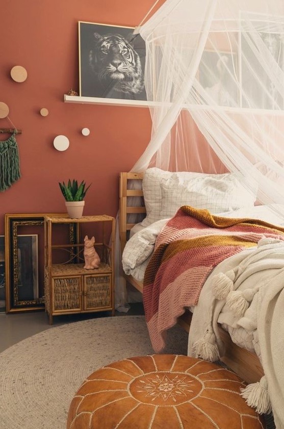 an eclectic bedroom with a terracotta accent wall, wooden and rattan furniture, neutral and bright bedding, a canopy and a gallery wall on a ledge