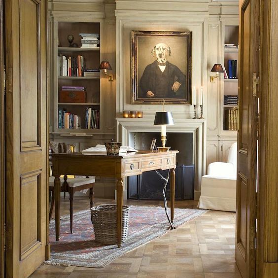 an exquisite vintage neutral home office with built-in bookshelves, a stained desk, creamy chairs, some artwork, a basket under the desk