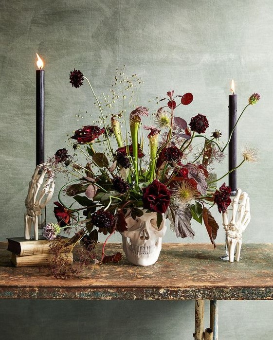 a skull vase with burgundy blooms, dark and usual leaves, greenery and black candles for Halloween