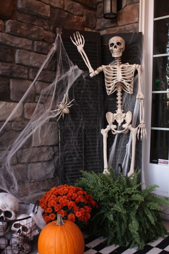 a Halloween porch styled with shutters, a skeleton, spiderweb, greenery and blooms, skulls and a bold pumpkin is a cool idea