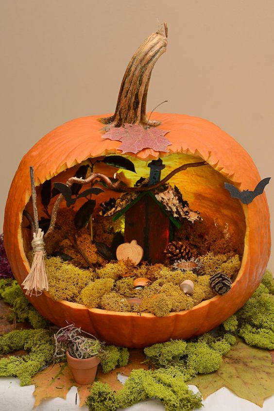a Halloween pumpkin diorama with moss, pumpkins, pinecones, a broom, bats and a small house with lights behind it is a great idea