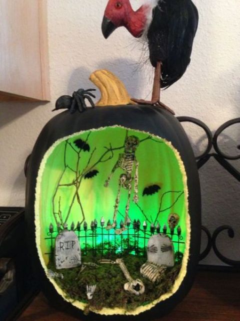 a black and green inside pumpkin wiht a graveyard, a fence, a skeleton, bats and branches is a gorgeous idea of a diorama