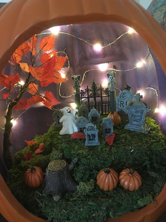 a graveyard pumpkin diorama with moss, mini pumpkins, tombstones, a ghost, lights and a fall tree is a great idea for Halloween