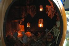 a moody Halloween diorama with a graveyard, lights, a black house and a skeleton plus little pumpkins is a bold idea