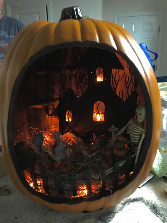 a moody Halloween diorama with a graveyard, lights, a black house and a skeleton plus little pumpkins is a bold idea