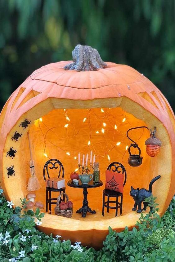 a stylish Halloween pumpkin diorama with lights, small black chairs and a table with candles, a kettle, a black cat and some black spiders