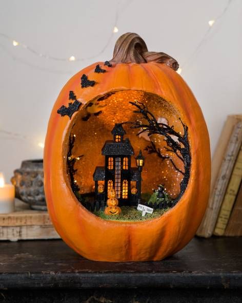 an orange pumpkin diorama with a black house, a tree, a stack of pumpkins and grass and bats on the side of the pumpkin