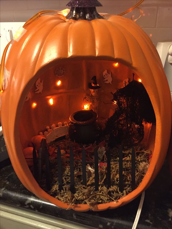 an orange pumpkin diorama with hay, a fence, lights, a cauldron, a skeleton in a witch hat and some lights