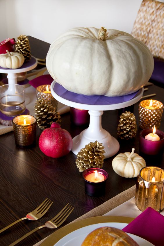 a bold Thanksgiving tablescape with gilded pinecones, a white pumpkin on a purple stand, fuchsia napkins and pomegranats, purple candleholders