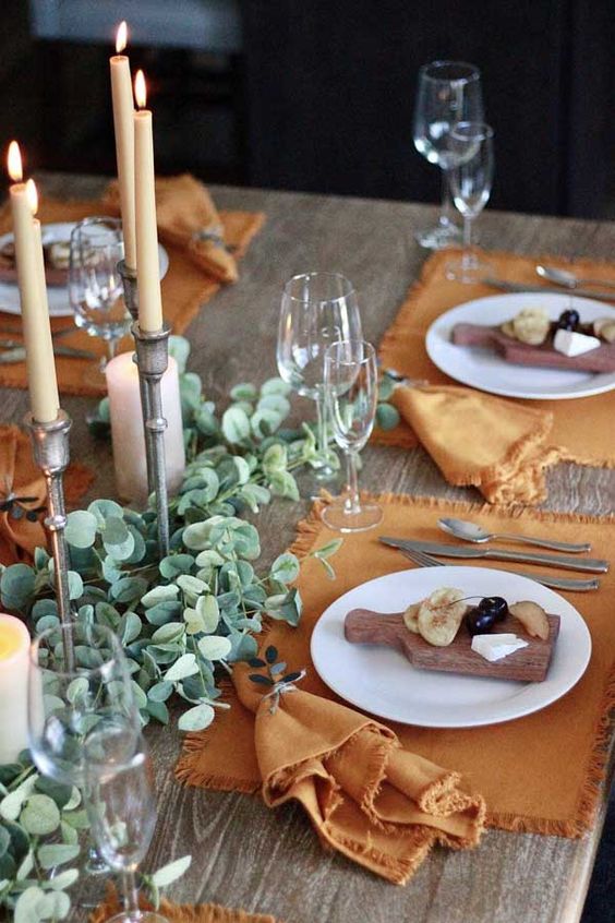 a beautiful Thanksgiving table setting with orange placemats and napkins, greenery table runners, candles and boards