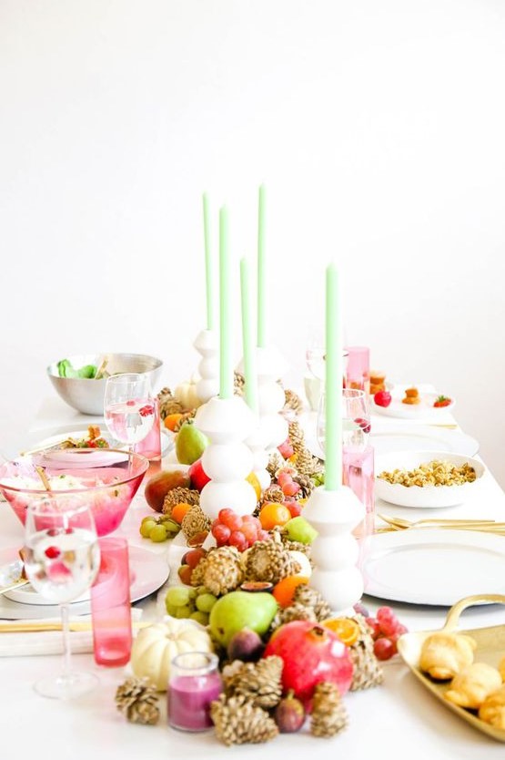 a bold Thanksgiving tablescape with pink glasses, green candles, bold fruit and candles on the table and some pinecones