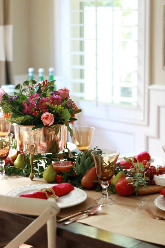 a bright Thanksgiving table setting with bold blooms, fruit, veggies and greenery, red napkins and gilded cutlery