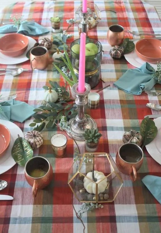 a bright Thanksgiving tablescape with a bold plaid tablecloth, greenery, pumpkins, pink candles, copper mugs, white and pink porcelain