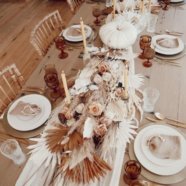 a gorgeous boho Thanksgiving tablescape with gold placemats and white porcelain, a lush table runner made of pampas grass, fronds, a white pumpkins, pink and white blooms