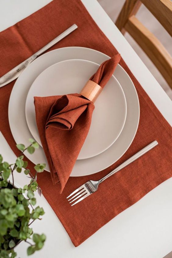 a stylish and laconic Thanksgiving place setting with a rust placemat and a napkin, white plates and a copper napkin ring