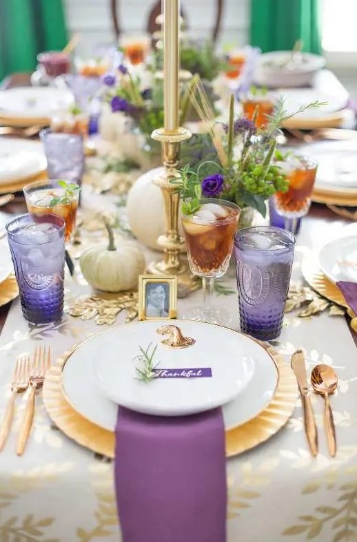 a refined gold and purple Thanksgiving tablescape with gold cutlery, chargers and candlesticks, white pumpkins, greenery, purple glasses and napkins