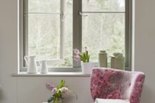 a chic cottage-inspired space with a green frame casement window, a green bench and a pink floral chair plus some blooms