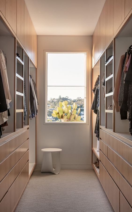 a contemporary narrow closet with stained wood drawers, cabinets and shelves and some railings for clothes plus a window for natural light