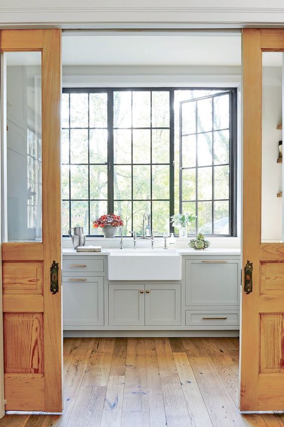 a farmhouse kitchen with grey shaker cabinets, white countertops, a large black frame casement window that lets a lot of light in