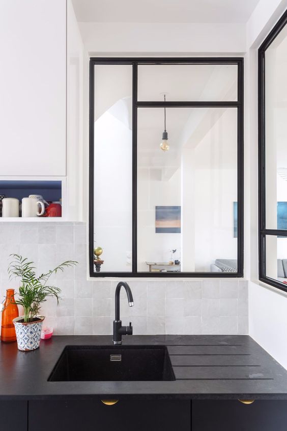 a kitchen with twon black framed windows is connected to the living room and gets a lot of light plus you can watch people cooking