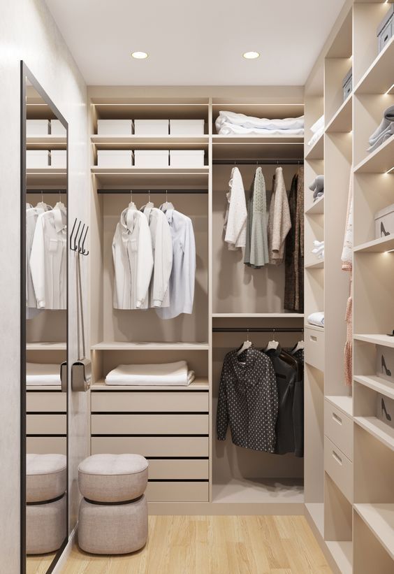 a narrow and small closet with neutral shelves, closets and lights and a large mirror plus a small pouf