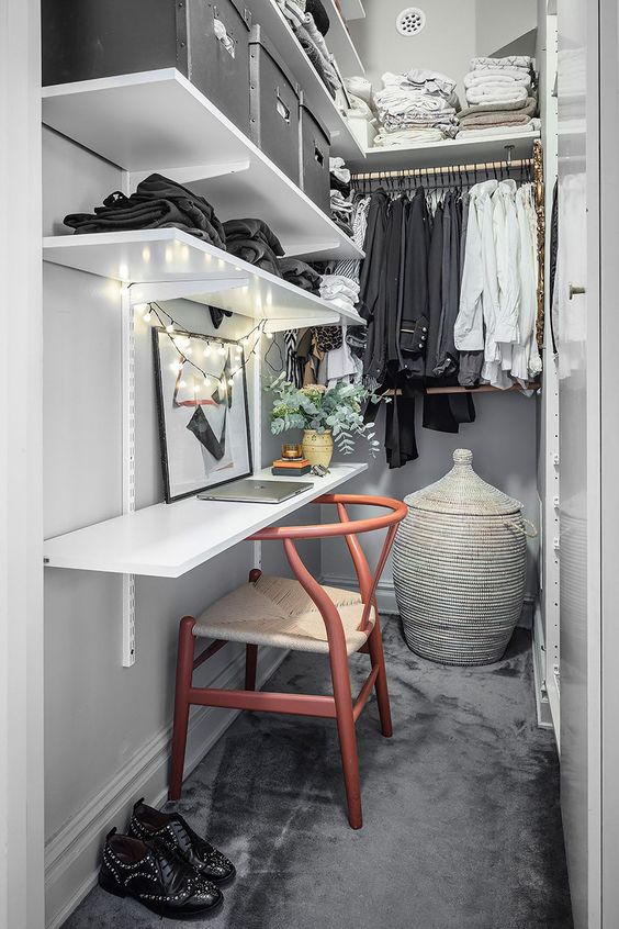 a narrow closet done with open shelves, railings, a shelf that is a desk, a chair, a basket with a lid and some lights