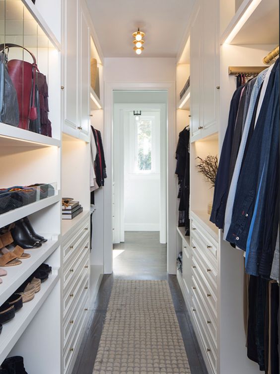 a narrow white walk in closet with shelves, open storage compartments, railings, lights and lit up shelves