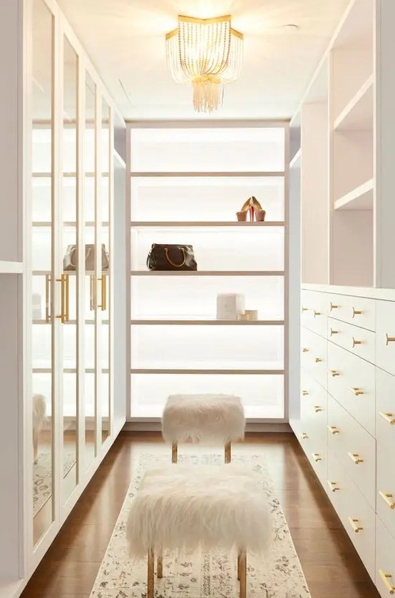 a neutral and very chic closet with drawers with glass doors, open shelves and drawers, open shelves and faux fur stools plus a crystal chandelier