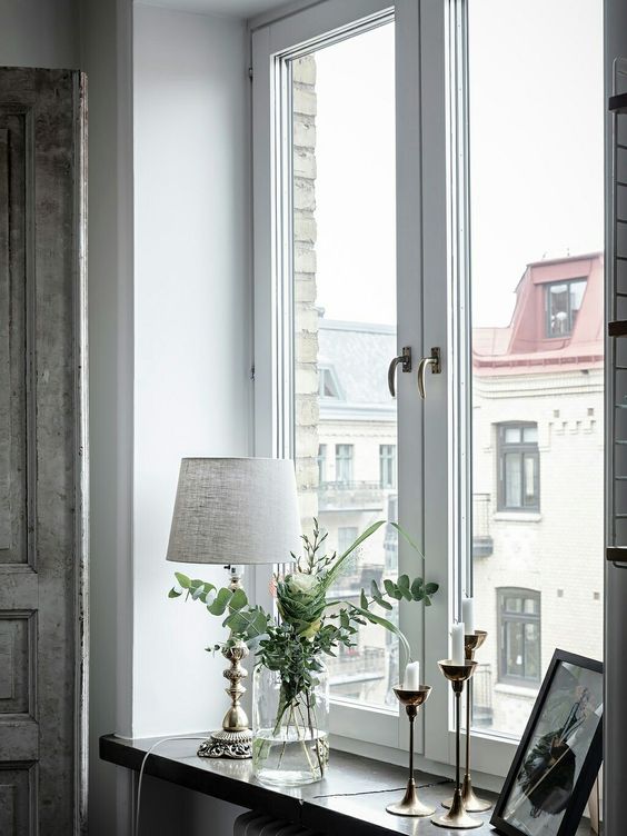 a neutral space with a stylish casement window, a windowsill decorated with candles, a lamp, some greenery and artwork
