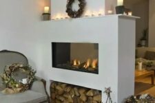 a sleek white fireplace with firewood storage, candles on the mantel and a wreath with pinecones for winter