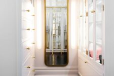 a small and elegant narrow closet done with white storage units, with glass doors, drawers and a curved mirror in a gilded frame