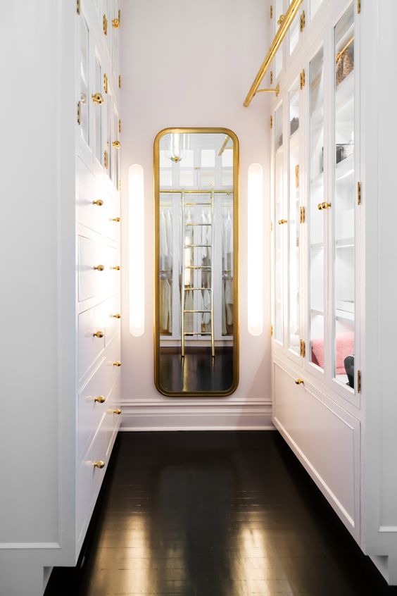 a small and elegant narrow closet done with white storage units, with glass doors, drawers and a curved mirror in a gilded frame