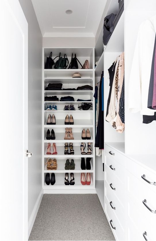 a small and narrow white walk-in closet with holders for clothes, open shelves for shoes, drawers for smaller stuff is cool and organized