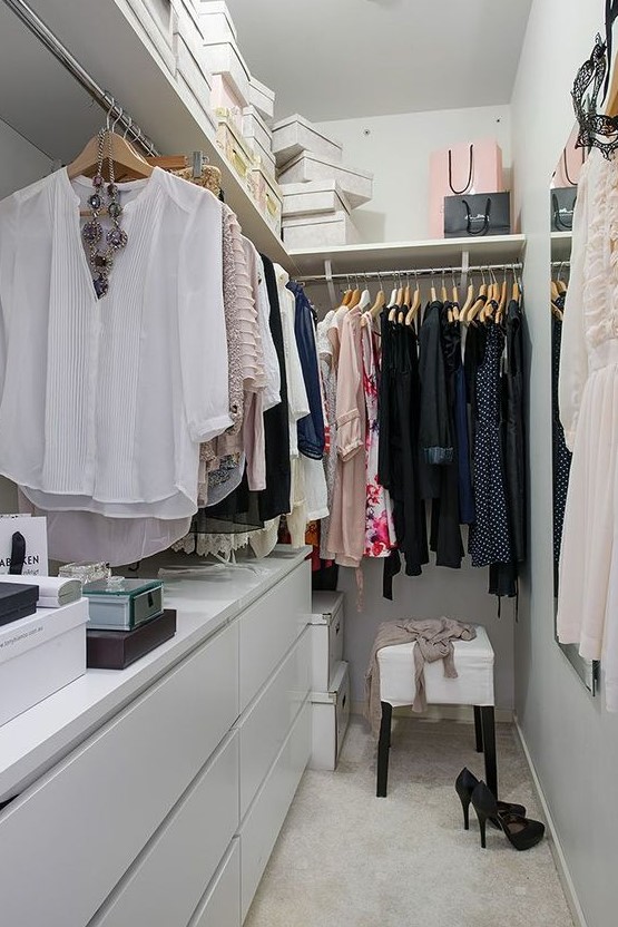 a small contemporary closet in white, with a large open shelf with boxes, dressers, holders with hangers and a stool