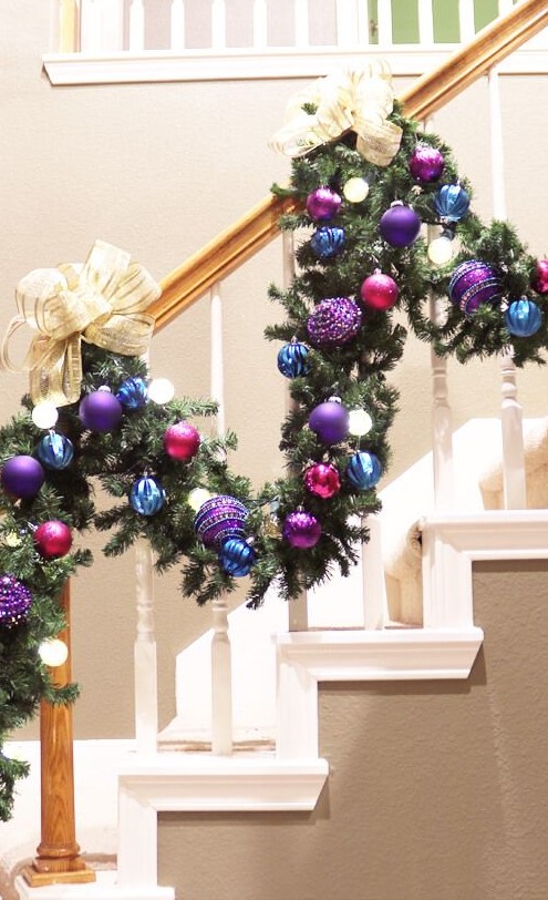 a staircase garland with blue, purple and fuchsia ornaments and large neutral bows and lights is amazing for a bright touch