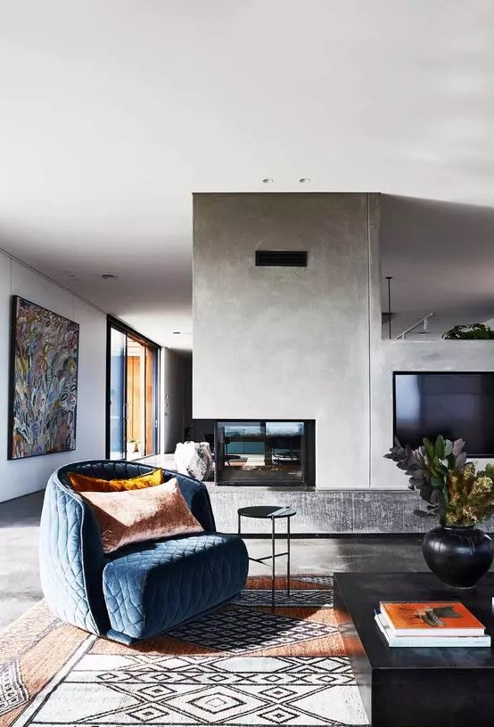 a stylish mid-century modern living room with a concrete double-sided fireplace, a blue quilted chair, a printed rug and a bold artwork
