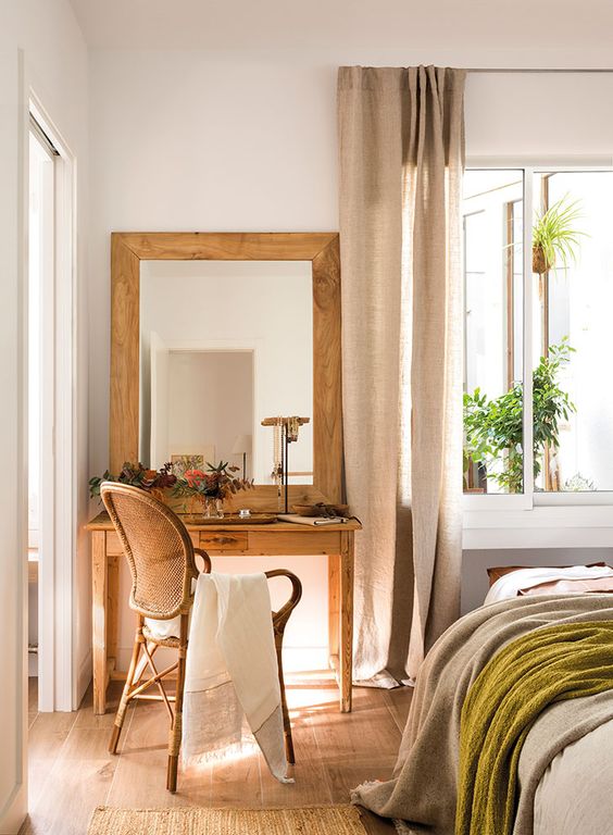 a nostalgic bedroom in neutrals, with a wooden vanity and a mirror in a wooden frame, a vintage chair and a bed with neutral bedding