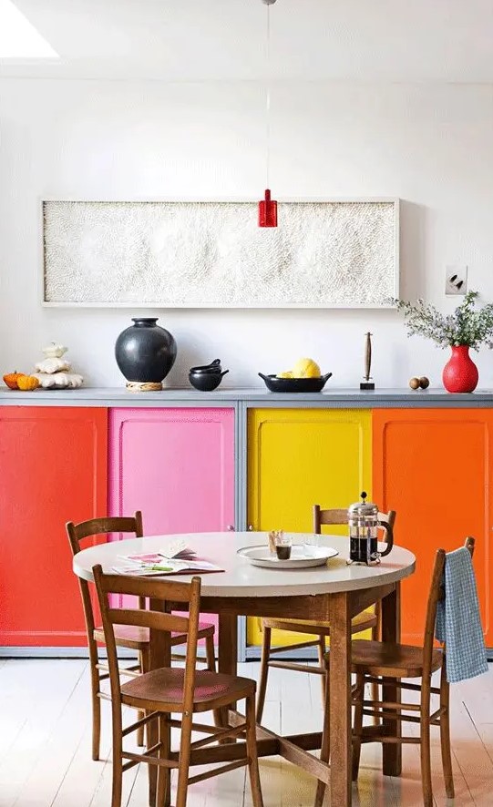a colorful kitchen with bold doors, and a round table, wooden chairs and a pretty textural artwork and red touches
