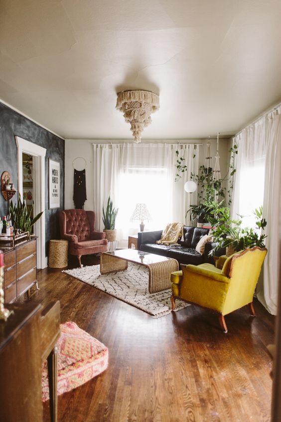 a nostalgic living room with a black limewash wall, a burgundy and yellow chair, a black leather sofa, a rattan table and some boho decor