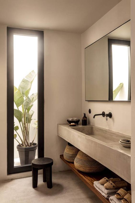 a concrete Japandi bathroom with a large mirror, a built-in concrete vanity and a shelf, a vertical window plus a black stool