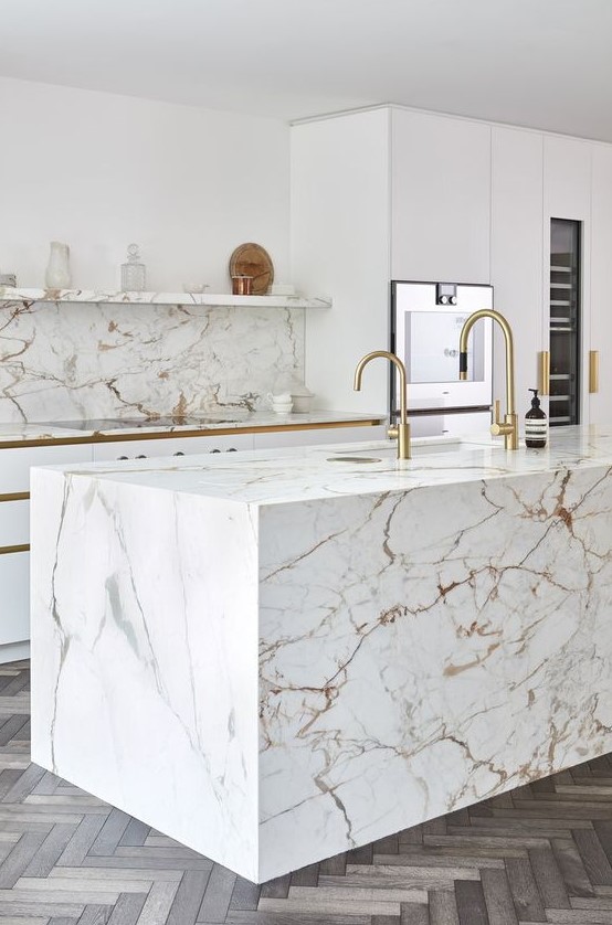 a modern luxurious kitchen with white cabinets, a white marble kitchen island and a backsplash plus gold fixtures