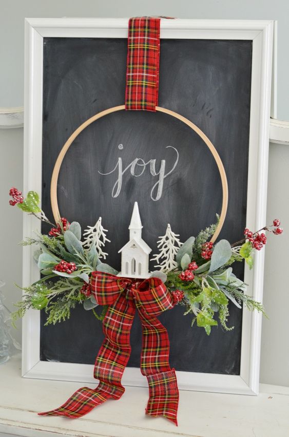 a small and cool embroidery hoop Christmas wreath with greenery, berries, a red plaid bow, a church and white faux trees