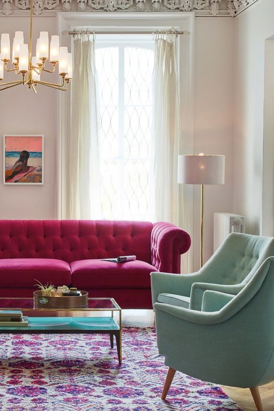 a beautiful living room with a magenta sofa, pastel green chairs, a chic chandelier, a bold boho rug, a low table and neutral curtains