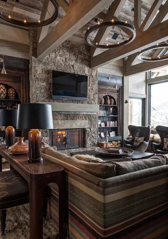 a chalet living room with a fireplace clad with stone, striped seating furniture, metal chandeliers and table lamps