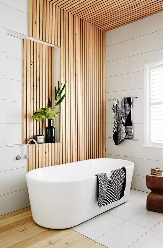a contemporary neutral bathroom with white large scale tiles, wooden slats, a niche for storage, an oval tub and a wooden stool