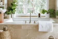 30 a gorgeous neutral spa bathroom clad with limestone tiles, with a tub clad with them, built-in shelves and potted greenery, a woven pouf and a bag