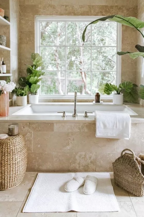 a gorgeous neutral spa bathroom clad with limestone tiles, with a tub clad with them, built-in shelves and potted greenery, a woven pouf and a bag