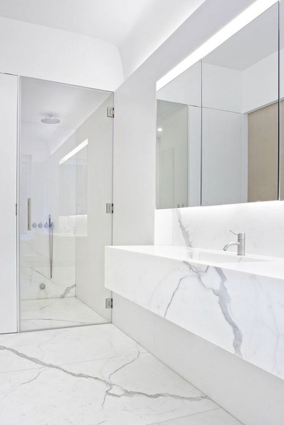 a clean minimalist space done with white marble, a mirror with lights, a shower space and a marble vanity