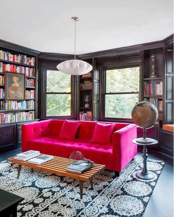 a moody and elegant black home library with lots of bookshelves, a magenta sofa and a printed rug, a globe and a pendant lamp