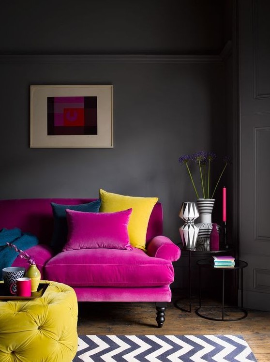 a moody living room with grey walls, a magenta sofa, a yellow tufted ottoman, a printed rug, chic vases and candles